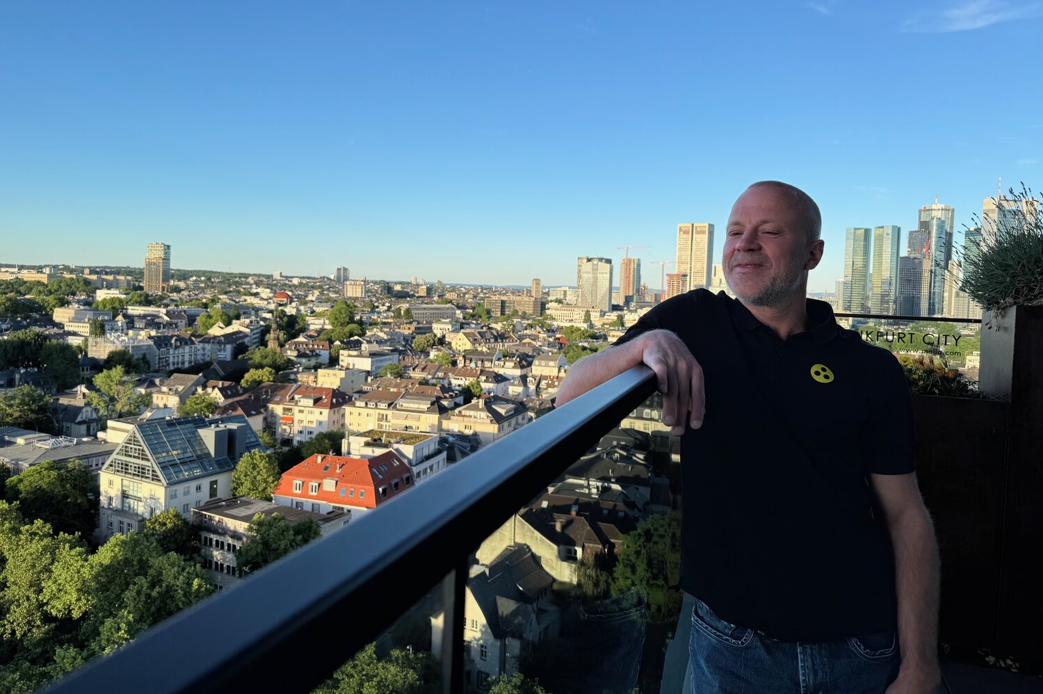 Florian Schwab auf einer Dachterrasse in Frankfurt, an eine Reling gelehnt, im Hintergrund die Skyline mit Hochhäusern bei blauem Himmel.