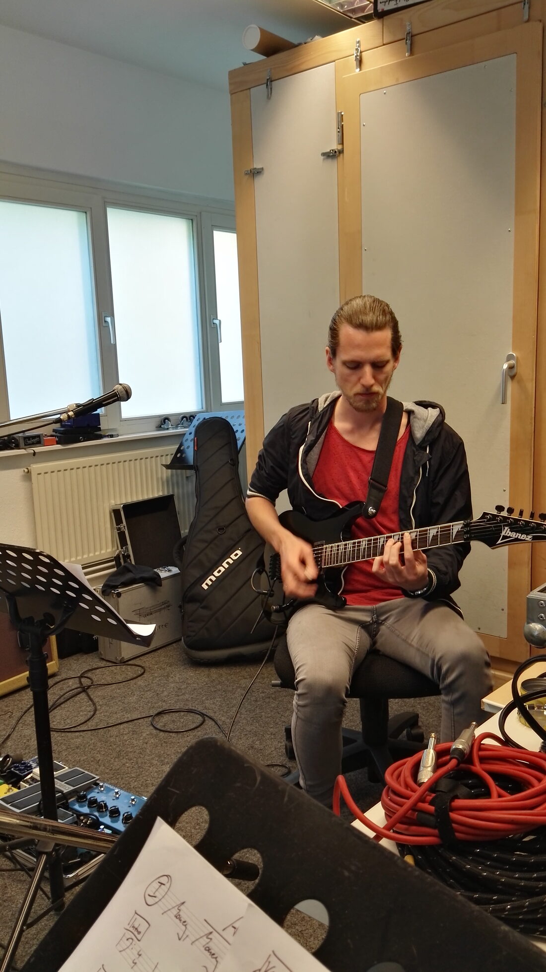 Florian Schwab sitzt in seinem Studio und spielt E-Gitarre, konzentriert, umgeben von Mikrofonstativ, Notenständer, Gitarrenkoffer und Kabeln.