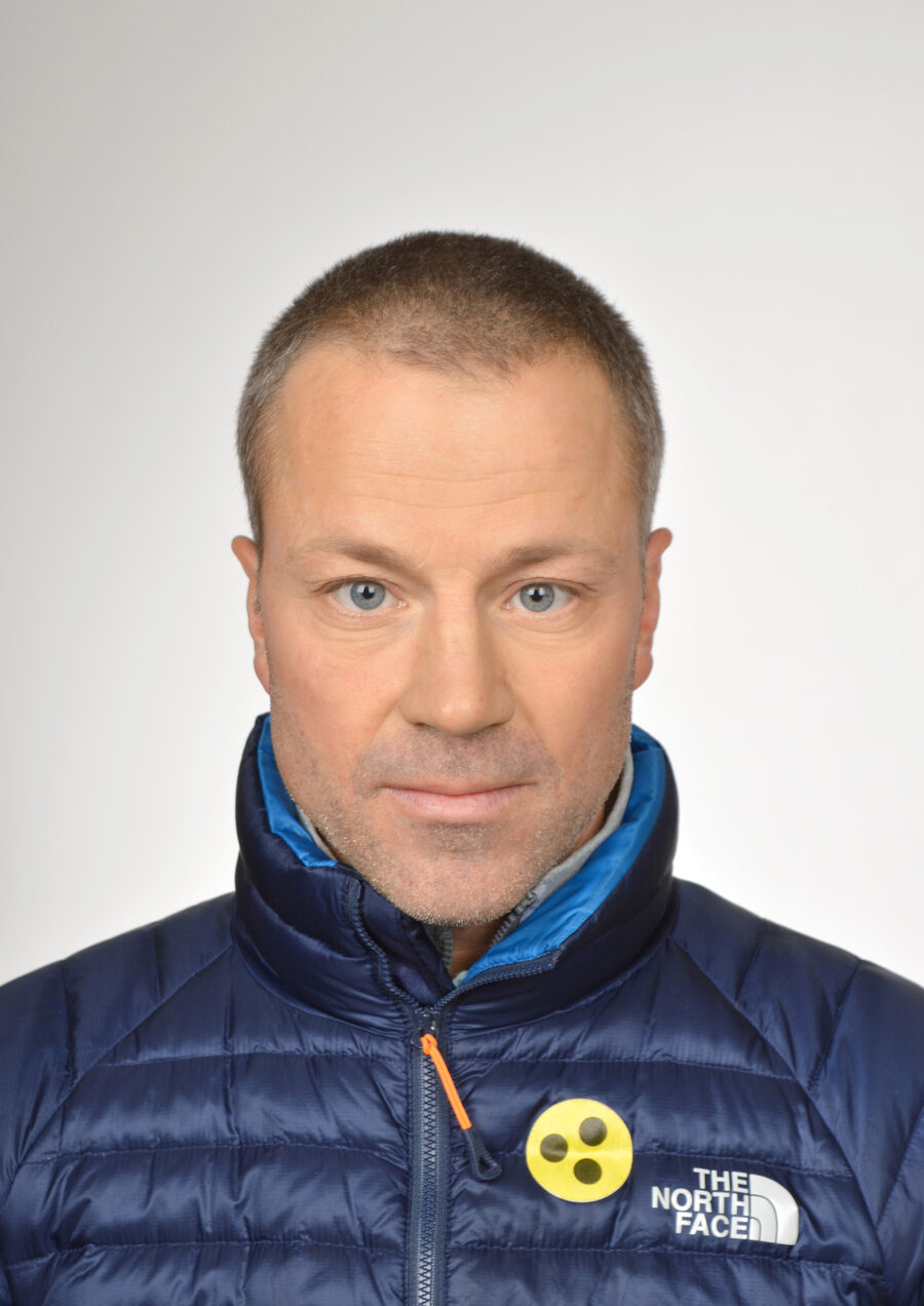 Porträt von Florian Schwab vor hellem Hintergrund, in dunkelblauer Steppjacke, mit dem gelben Abzeichen für blinde und sehbehinderte Menschen.